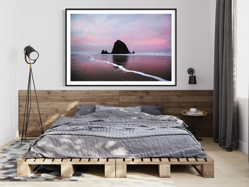 May include: A framed photograph of a large rock formation on a beach at sunset. The sky is a vibrant pink and orange, and the water is reflecting the colors of the sky. The rock formation is silhouetted against the sky.