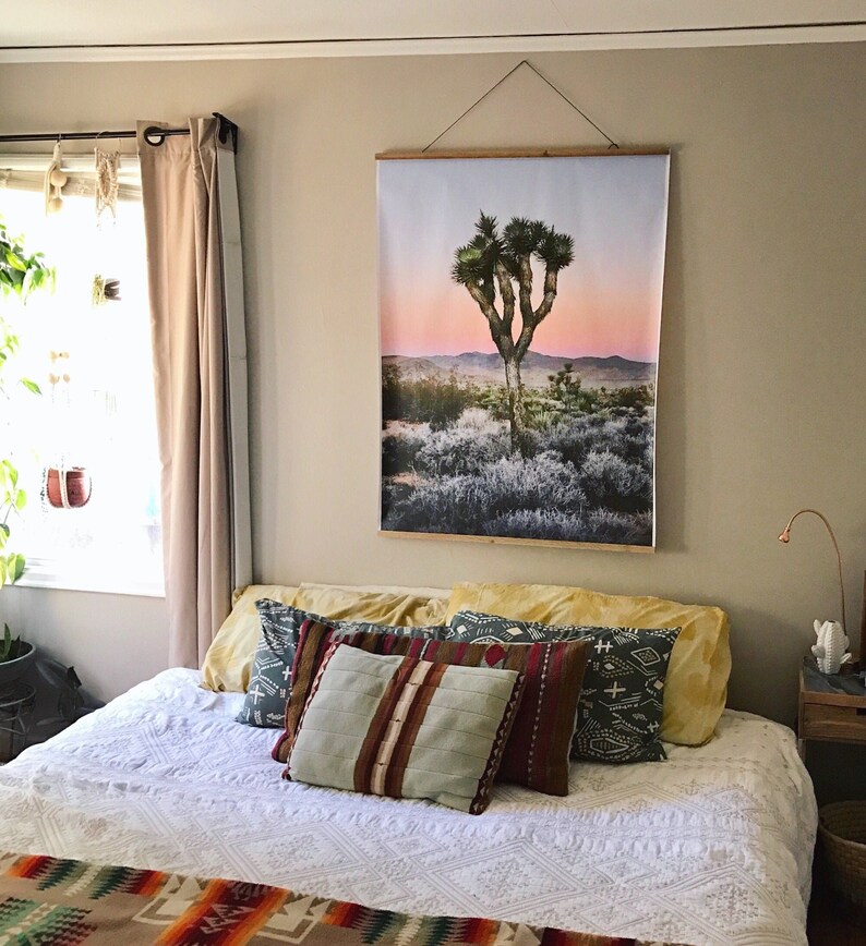 May include: A white bed with a white crocheted bedspread and a variety of patterned throw pillows. A large framed print of a Joshua tree in a desert landscape hangs on the wall above the bed.