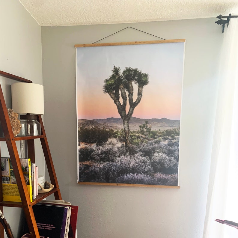 May include: A framed print of a Joshua Tree in a desert landscape. The tree is silhouetted against a pink and orange sunset sky. The print is mounted on a wooden frame.