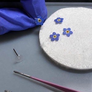 May include: Small blue forget-me-not flowers with yellow centers, preserved and encased in clear resin. The set includes individual flower components, a clear earring post with a flat pad, and a pair of pink metal tweezers for assembly.