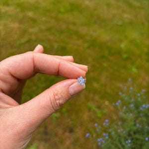 May include: A small stud earring shaped like a five-petaled flower with light blue petals and a yellow center. The earring is held between two fingers, showcasing its delicate size and floral design against a blurred natural green background.