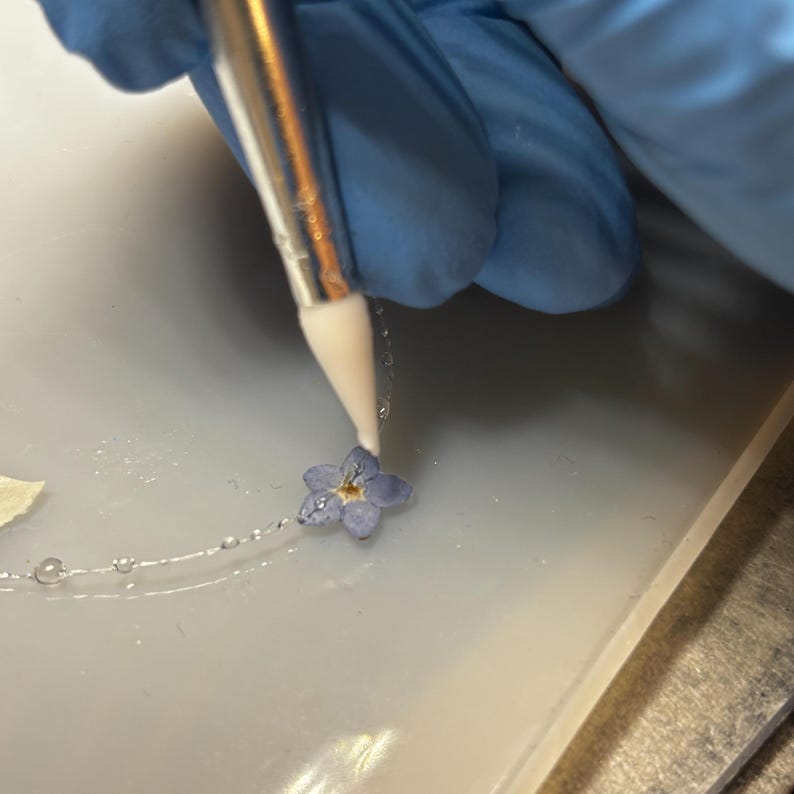 May include: A delicate silver chain necklace featuring a small, pressed light blue forget-me-not flower. A white-tipped applicator tool is positioned directly over the center of the flower, which has five petals and a yellow-brown center.