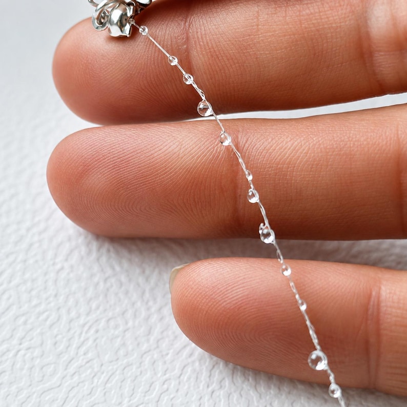 May include: A delicate, thin, transparent nylon thread featuring small, clear, spherical beads spaced at intervals along its length, attached to a metallic silver-toned component at the top end, creating a subtle, minimalist, and lightweight jewelry element.