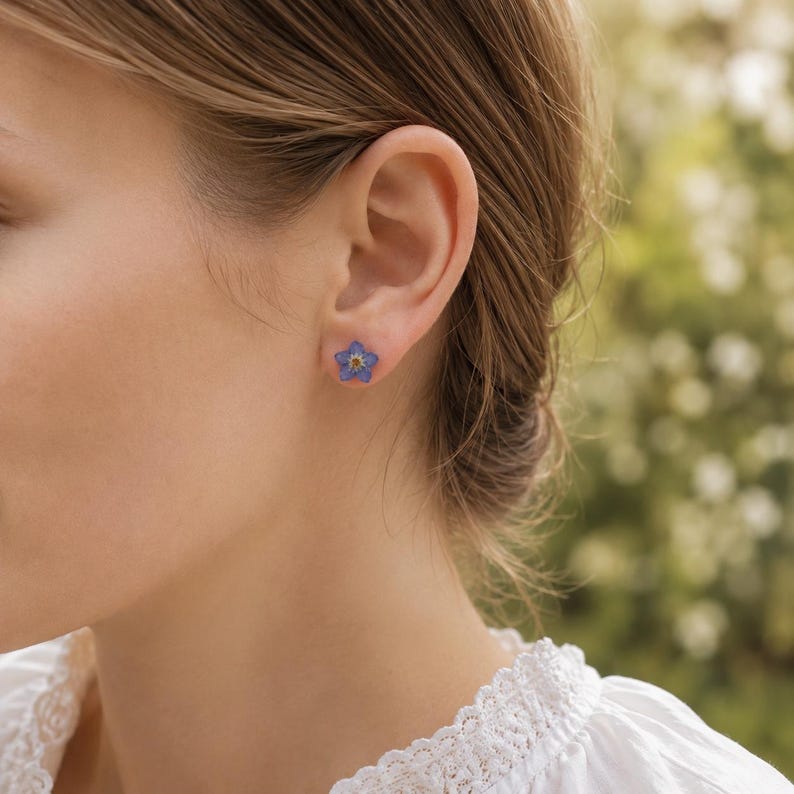May include: A small stud earring shaped like a five-petaled flower. The petals are a vibrant blue color with a yellow center, resembling a forget-me-not blossom. The earring is secured to the earlobe with a simple metal post and backing.