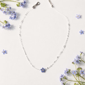 May include: A delicate necklace featuring a clear, thin cord adorned with small, reflective silver beads and a single, pressed blue forget-me-not flower pendant at the center. The necklace includes a silver-toned lobster clasp and an extender chain.