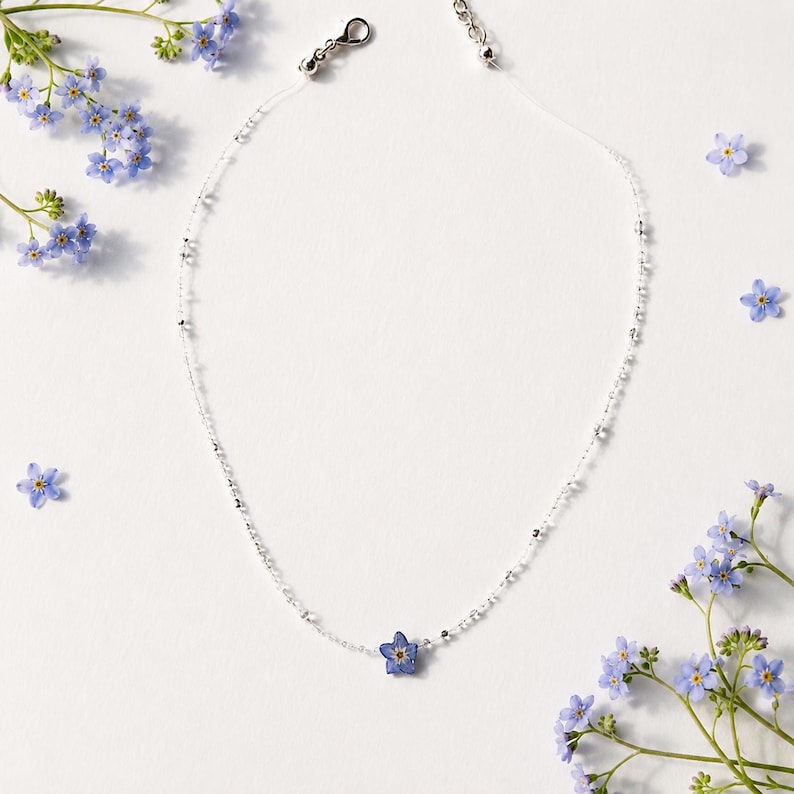 May include: A delicate necklace featuring a clear, thin cord adorned with small, reflective silver beads and a single, pressed blue forget-me-not flower pendant at the center. The necklace includes a silver-toned lobster clasp and an extender chain.