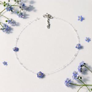 May include: A delicate silver chain necklace featuring three small, blue forget-me-not flowers spaced evenly along the strand. The necklace includes a silver clasp and an adjustable chain link extension, accented by loose blue flowers nearby.