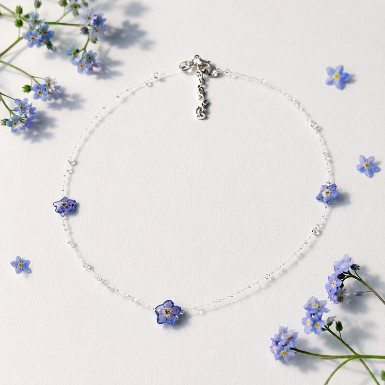May include: A delicate silver chain necklace featuring three small, blue forget-me-not flowers spaced evenly along the strand. The necklace includes a silver clasp and an adjustable chain link extension, accented by loose blue flowers nearby.