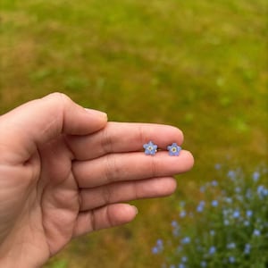 May include: A pair of small stud earrings shaped like five-petaled flowers. Each earring features light blue petals with a small yellow center. The earrings are positioned on a finger, showcasing their delicate size and floral design.