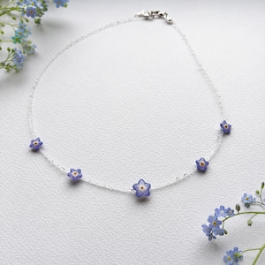 May include: A delicate necklace featuring a thin, clear, transparent cord adorned with five small, pressed blue forget-me-not flowers with yellow centers, spaced evenly along the strand, finished with a silver-toned metal lobster claw clasp.