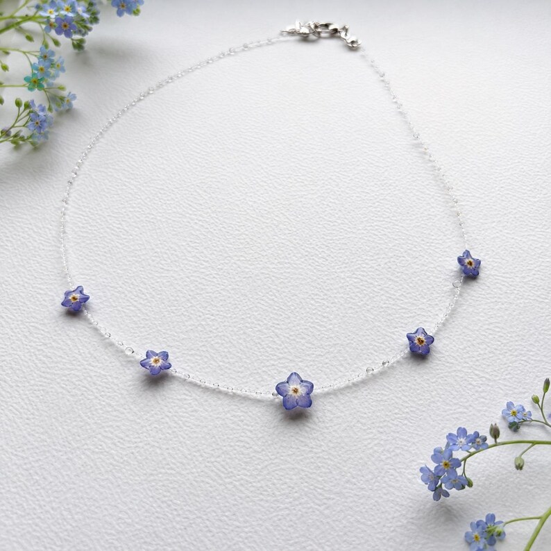 May include: A delicate necklace featuring a thin, clear, transparent cord adorned with five small, pressed blue forget-me-not flowers with yellow centers, spaced evenly along the strand, finished with a silver-toned metal lobster claw clasp.
