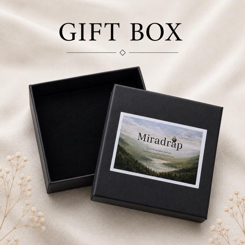 May include: A black square gift box with an open lid, featuring a rectangular label on the top surface. The label displays a landscape illustration of a valley with the text Miradrap, From Norwegian nature, Captured and preserved forever.