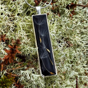 Puede incluir: Collar con colgante rectangular y cadena plateada. El colgante presenta un fondo negro con delicadas semillas blancas de diente de león y pequeñas cabezas de semillas doradas encapsuladas en resina transparente. El colgante está enmarcado en plata.