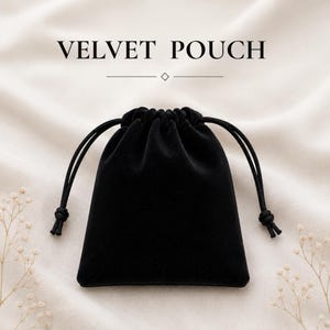 May include: A black velvet pouch with a drawstring closure featuring two black cords with knotted ends. The text VELVET POUCH is printed in black above the pouch, accompanied by a horizontal line and a small diamond-shaped icon in the center.