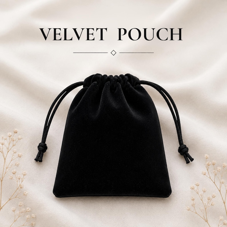 May include: A black velvet pouch with a drawstring closure featuring two black cords with knotted ends. The text VELVET POUCH is printed in black above the pouch, accompanied by a horizontal line and a small diamond-shaped icon in the center.
