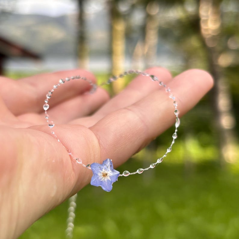 May include: A delicate necklace featuring a single, small, light blue five-petaled flower pendant with a yellow center, suspended on a thin, clear, translucent filament chain adorned with tiny, spaced-out, clear, bead-like accents along the length.
