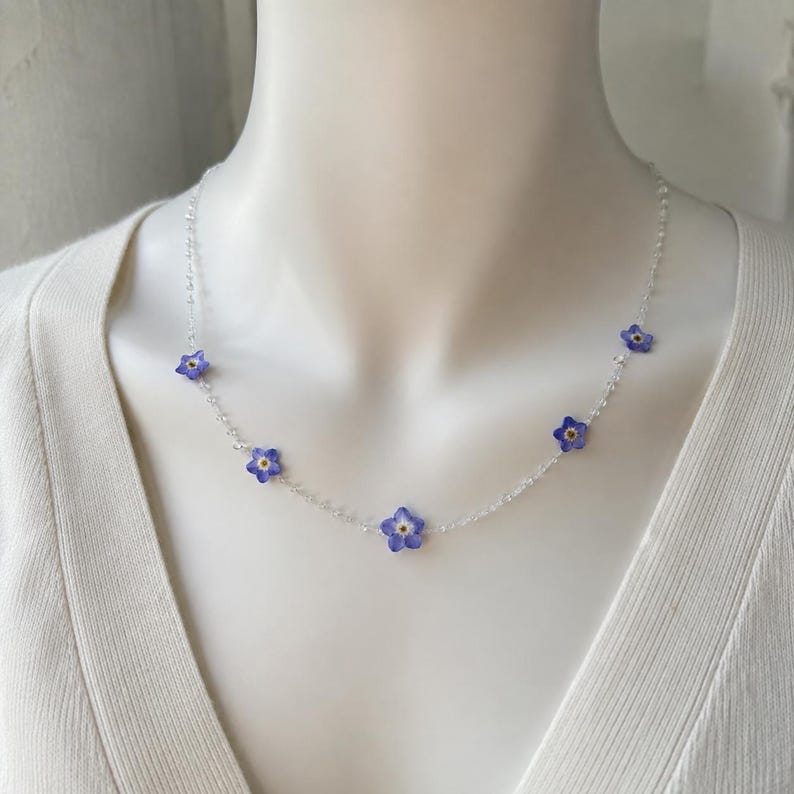 May include: A delicate necklace featuring a thin, clear, beaded chain interspersed with five small, five-petaled blue forget-me-not flowers. Each flower has a tiny yellow center, and the necklace rests against a plain white fabric surface.