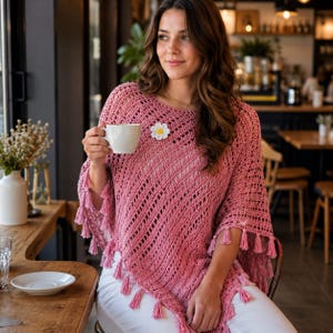 Könnte beinhalten: Ein rosafarbener Häkelponcho mit einem durchbrochenen diagonalen Maschenmuster, einem Rundhalsausschnitt und einem Saum, der mit kleinen passenden rosa Quasten verziert ist. Eine weiß-gelbe gänseblümchenförmige Brosche ist am Oberkörper befestigt.