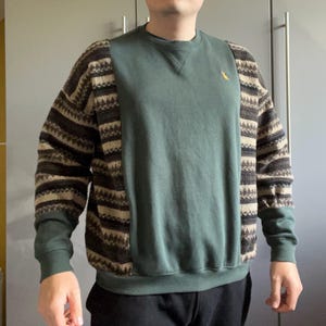 May include: Green sweatshirt with patterned sleeves. The sleeves feature a brown, tan, and black geometric design. A small yellow bird is embroidered on the chest. The sweatshirt has a crew neck and ribbed cuffs.