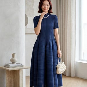 Puede incluir: Un vestido midi azul marino con cuello alto, mangas cortas y una textura plisada en toda la prenda. El vestido presenta un corpiño ajustado que se transforma en una falda acampanada y voluminosa. Se incluye un pequeño bolso blanco con asa de perlas.