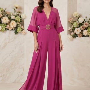 May include: A magenta jumpsuit featuring sheer elbow-length sleeves, a deep V-neckline, and wide-leg trousers with side slits. The garment includes a matching belt at the waist adorned with a large, circular gold-toned decorative buckle.