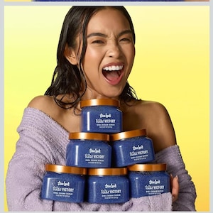 Puede incluir: Una pila de seis tarros azules de exfoliante de az&uacute;car de karit&eacute; con tapas doradas. Los tarros tienen el texto "True Luck Valet Victory Shea Sugar Scrub" impreso. El fondo es amarillo.