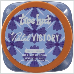 Puede incluir: Un tarro de exfoliante de az&uacute;car de karit&eacute; Tree Hut Violet Victory. La etiqueta presenta un fondo azul claro con patrones florales morados y las palabras "Violet Victory". El tarro tiene un borde naranja y el texto "Soothe + Soften".