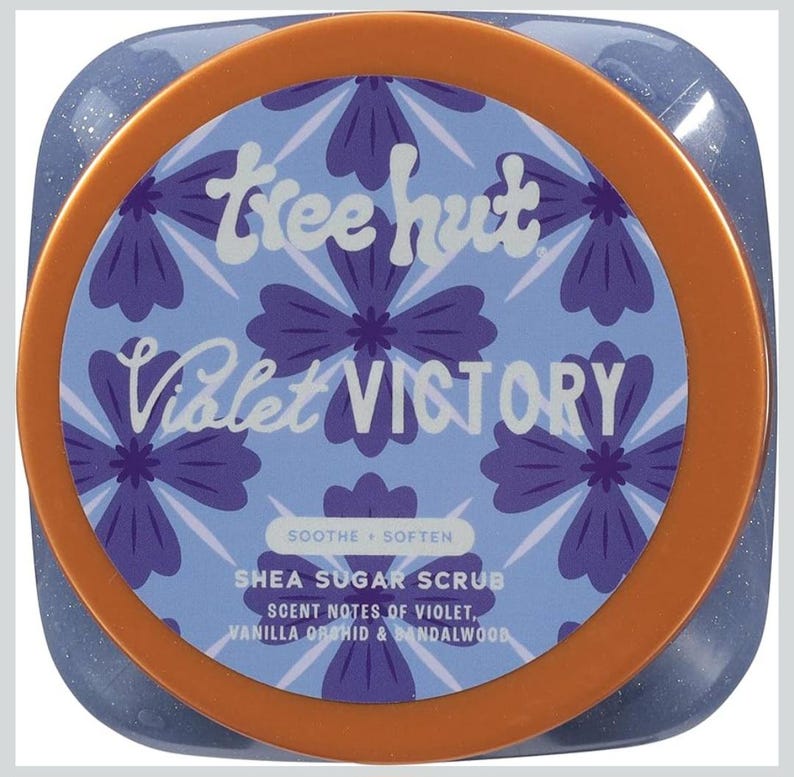 Puede incluir: Un tarro de exfoliante de az&uacute;car de karit&eacute; Tree Hut Violet Victory. La etiqueta presenta un fondo azul claro con patrones florales morados y las palabras "Violet Victory". El tarro tiene un borde naranja y el texto "Soothe + Soften".