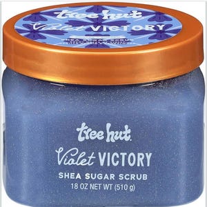 Puede incluir: Un tarro de exfoliante de az&uacute;car de karit&eacute; Tree Hut Violet Victory. El tarro tiene un cuerpo azul y una tapa de color cobre. La etiqueta dice "Violet Victory" y "Shea Sugar Scrub, 510 g". La tapa tiene un dise&ntilde;o floral.