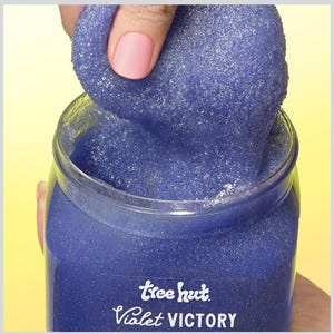Puede incluir: Un tarro de exfoliante corporal Tree Hut Violet Victory. El exfoliante es de color violeta intenso con una textura brillante. El tarro es transparente y la etiqueta presenta el nombre de la marca y del producto en escritura blanca.