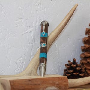 May include: A handcrafted pen with a wooden barrel, adorned with turquoise accents and silver-colored metal details. The pen is displayed on a wooden base, with a deer antler and pine cone in the background.