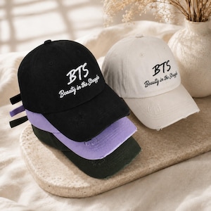 May include: A selection of baseball caps in black, purple, and green, with a cream-coloured cap in the background. Each cap features the embroidered text "BTS Beauty in the Struggle". The caps are displayed on a neutral-toned surface.