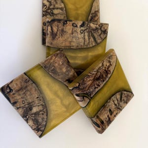 May include: Three square coasters made from a combination of natural wood with visible grain and dark knots, integrated with a translucent, shimmering mustard-yellow resin. The pieces are arranged in an overlapping stack on a plain white surface.