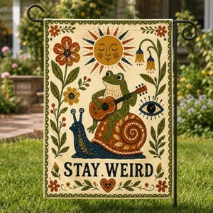 May include: A decorative garden flag with a quirky design. A green frog plays a guitar while sitting on a snail's shell. The flag features a sun, flowers, and the text "STAY WEIRD". The flag is bordered with a floral pattern.