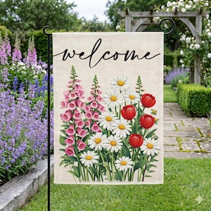 May include: A garden flag with the word "welcome" in black script. The flag features a floral design with pink foxgloves, white daisies, and red tulips. The flag is displayed on a black metal stand in a garden setting.