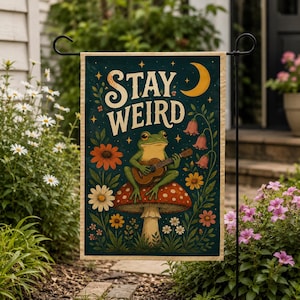May include: A rectangular garden flag featuring a dark teal background with the text STAY WEIRD in cream letters. A green frog sits on a red and white spotted mushroom playing a brown acoustic guitar, surrounded by colourful flowers and a yellow crescent moon.