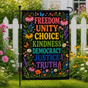 May include: A black garden flag featuring the words "FREEDOM UNITY CHOICE KINDNESS DEMOCRACY JUSTICE TRUTH" in rainbow colours. The flag is adorned with flowers and butterflies. It is displayed on a black metal stand in a garden with a white picket fence.