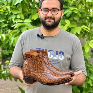 Puede incluir: Un par de botines marrones con un patrón de textura de piel de cocodrilo, punta puntiaguda y tacón apilado de color marrón oscuro. Las botas tienen un acabado pulido y una silueta estructurada con costuras visibles a lo largo de la suela.