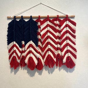 May include: A macrame wall hanging depicting the American flag. The artwork features a wooden dowel and a white cord for hanging. The design incorporates blue, red, and white knotted yarn, creating a textured, patriotic display.