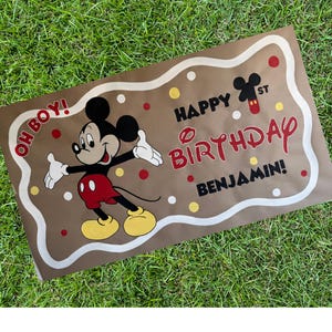 May include: A brown banner featuring Mickey Mouse with the text "Happy Birthday Benjamin!" in red and black. The banner has a white border and red, white, and yellow polka dots. Mickey Mouse is wearing red shorts and yellow shoes.