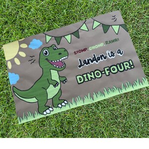 May include: A brown rectangular sign with a green dinosaur illustration and the text "Landon is a DINO-FOUR!". The sign also features a sun, clouds, and the words "STOMP. CHOMP. RAWR!" in a playful font. The sign is decorated with green bunting and grass.