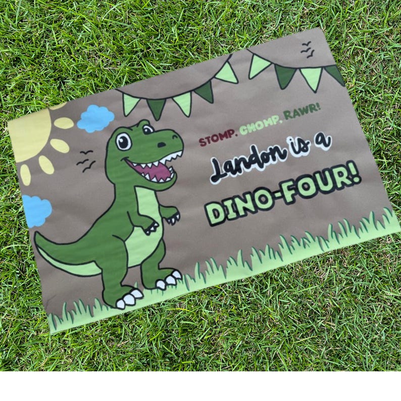 May include: A brown rectangular sign with a green dinosaur illustration and the text "Landon is a DINO-FOUR!". The sign also features a sun, clouds, and the words "STOMP. CHOMP. RAWR!" in a playful font. The sign is decorated with green bunting and grass.