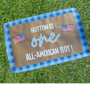 May include: A brown rectangular banner with blue gingham border. The banner reads "HUTTON IS one ALL-AMERICAN BOY!" in white and light blue script. Two American flags are on the banner.
