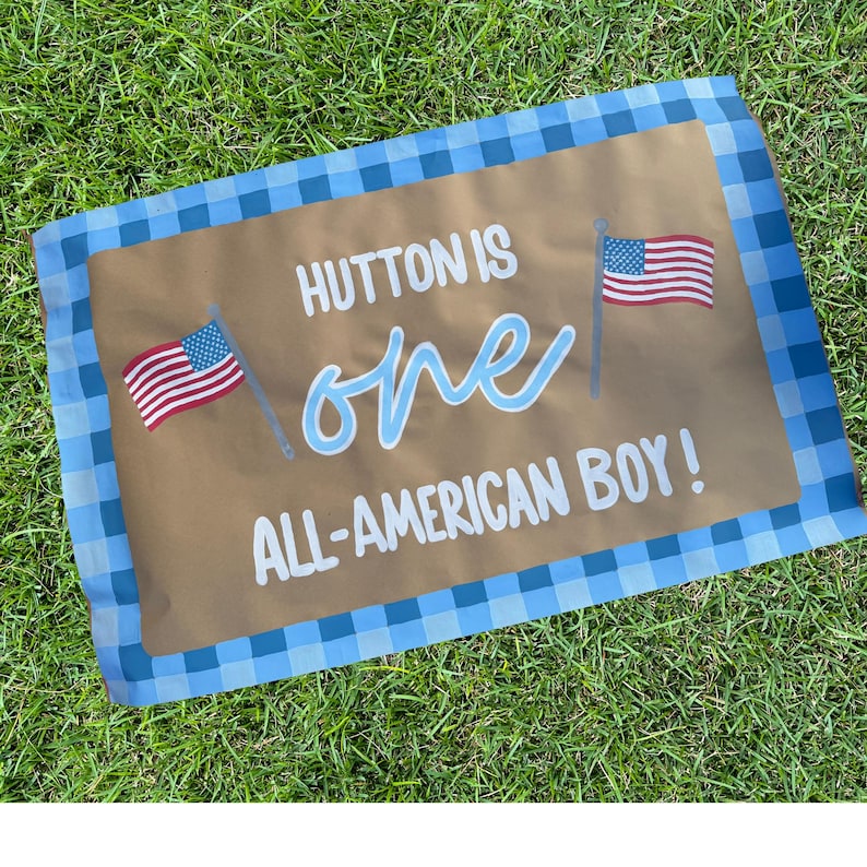 May include: A brown rectangular banner with blue gingham border. The banner reads "HUTTON IS one ALL-AMERICAN BOY!" in white and light blue script. Two American flags are on the banner.