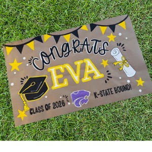 May include: A brown graduation sign with the words "Congrats EVA" in black and yellow. The sign includes a graduation cap, a diploma, stars, and the text "CLASS OF 2026" and "K-STATE BOUND!" on a green grass background.