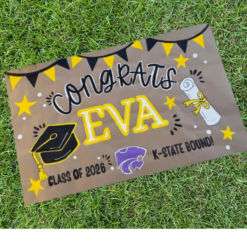 May include: A brown graduation sign with the words "Congrats EVA" in black and yellow. The sign includes a graduation cap, a diploma, stars, and the text "CLASS OF 2026" and "K-STATE BOUND!" on a green grass background.