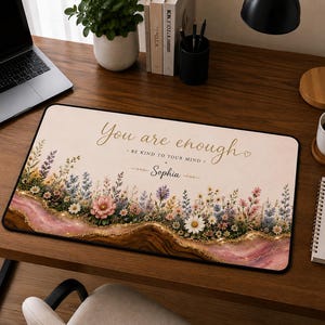 May include: A rectangular desk mat featuring a cream-colored background with a floral border of wildflowers in pink, blue, and white. The text reads "You are enough", "BE KIND TO YOUR MIND", and the name "Sophia" in gold script and print.