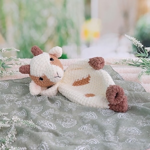 May include: A crochet security blanket featuring a plush goat head with white and tan yarn, dark brown ears, and small horns. The attached blanket body is made of soft white yarn with tan patches and dark brown accents at the corners.