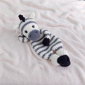 May include: A handmade crochet zebra plush toy with white and gray stripes, black hooves, and a dark gray muzzle. The stuffed animal is lying on a soft, pink blanket. The zebra has small black ears and a black mane.