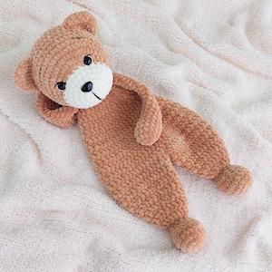 May include: A handmade, light brown crochet teddy bear with a white muzzle and black eyes. The bear is lying on a soft, light pink blanket. The bear's arms and legs are elongated, and it has a small, round tail.